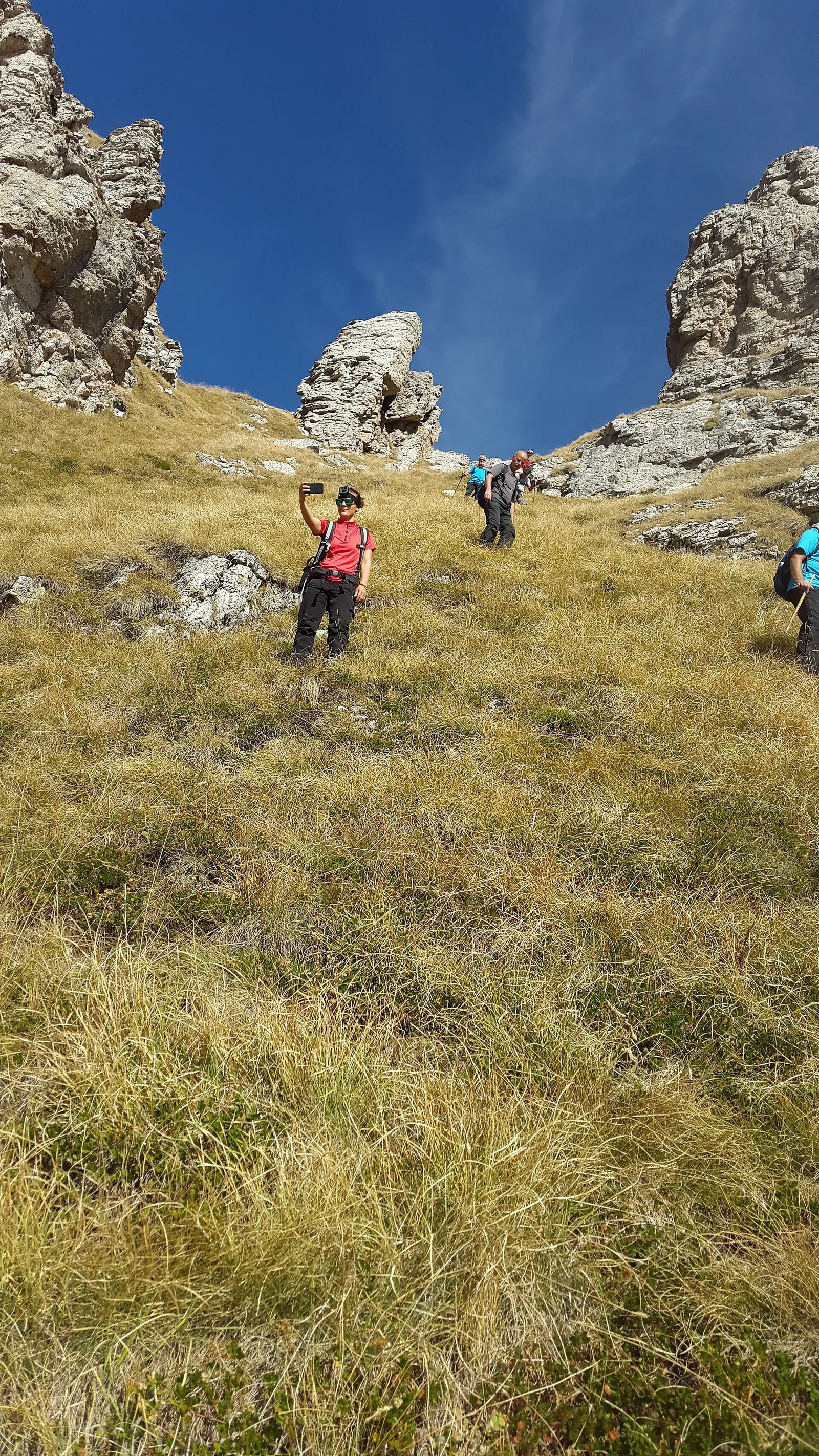 Hikers descending a grassy slope between limestone crags in the Albanian Alps under a clear blue sky