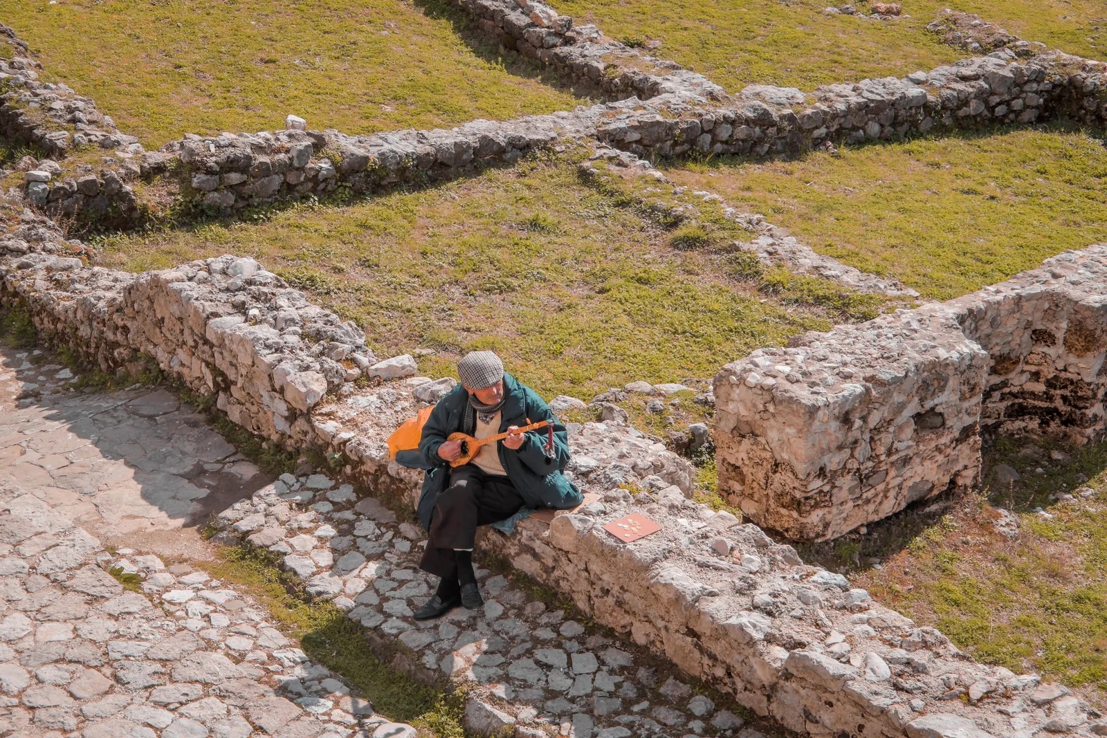 An Albanian musician playing a cifteli among old stone ruins on a grassy hillside