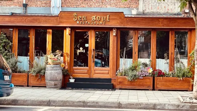 A wooden front with a carved sign, flower boxes along it and a barrel by the glass doors