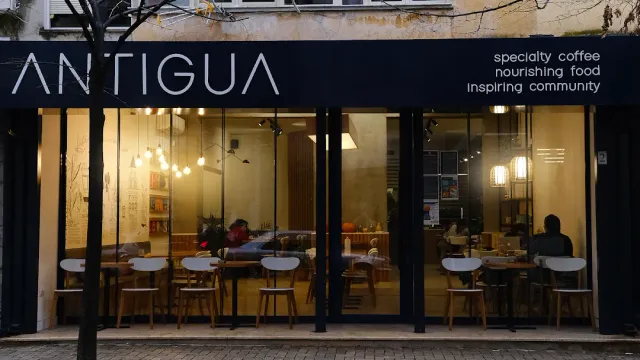 A cafe glass front with a dark sign, hanging bulbs and wooden tables inside