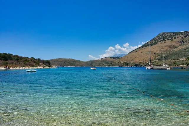 Shallow clear water over pebbles, anchored boats and dry hills across the bay