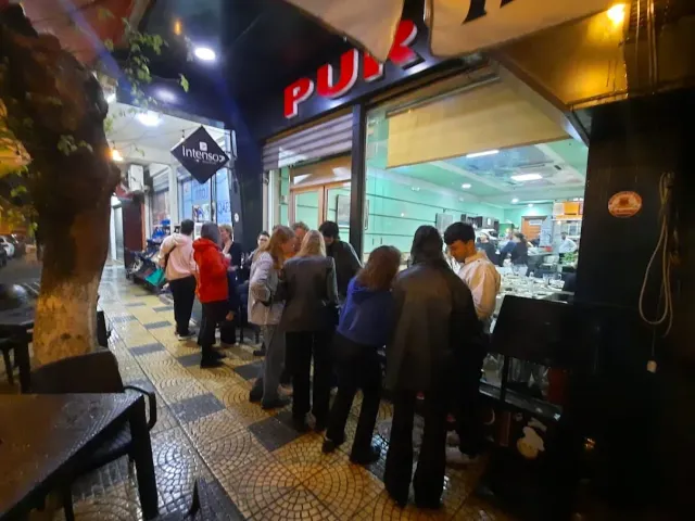 A queue of people at night at a lit street counter, a red sign and a paved pavement