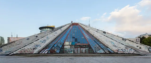 A sloping concrete pyramid shaped building with blue glass panels against a bright sky