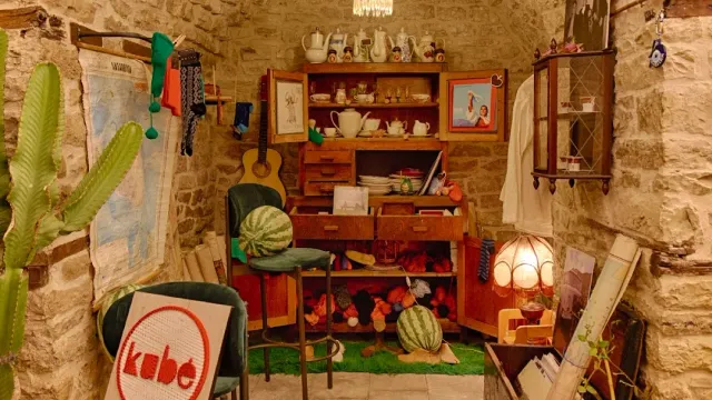 A small stone room with a wooden cupboard, china ware, a guitar, watermelons and a cactus in the corner
