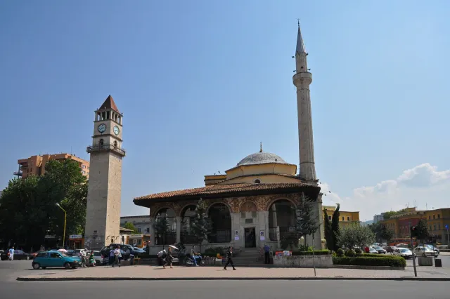A stone mosque with a dome and minaret beside a tall clock tower on a city square