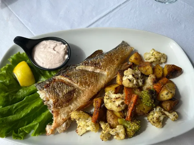 A whole grilled fish on a white plate beside roasted vegetables, a lemon wedge and white sauce