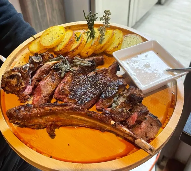 A wooden tray with roasted meat on the bone, potato slices and a bowl of sauce