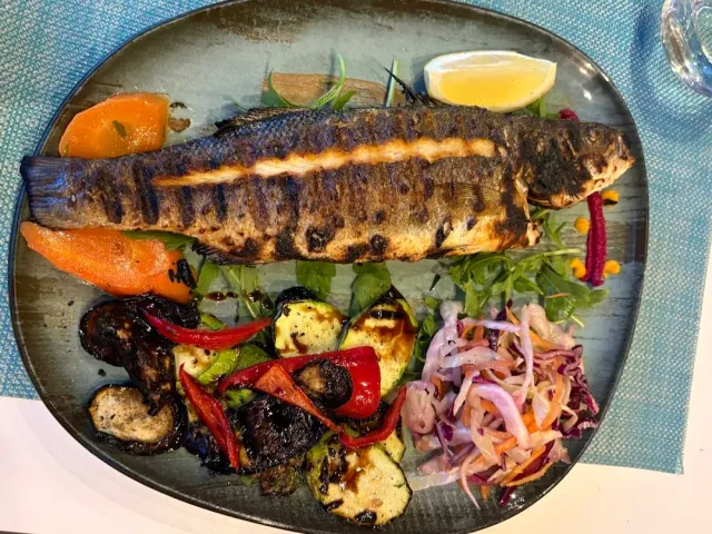 A whole grilled fish with roasted vegetables, cabbage salad and a lemon wedge on a plate