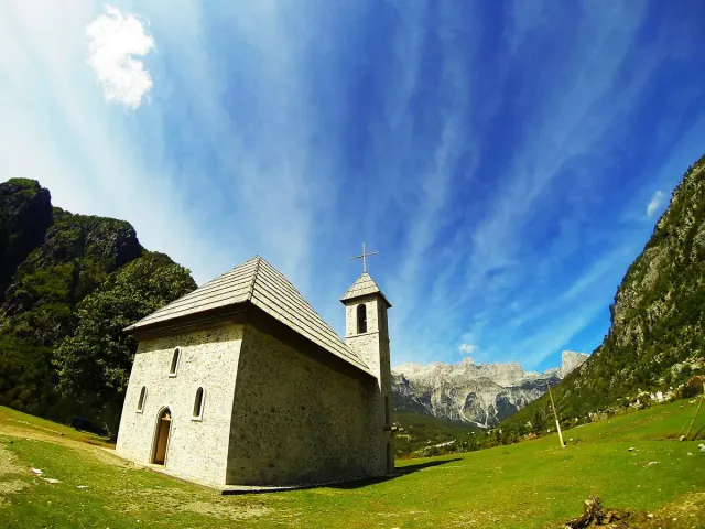 A small stone church with a tiled roof in a green meadow beneath rocky peaks