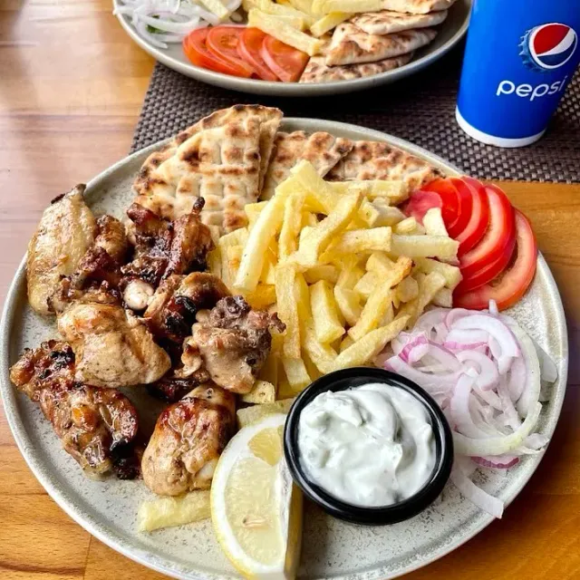 A plate with grilled chicken pieces, fries, pita, tomato, red onion and yogurt sauce