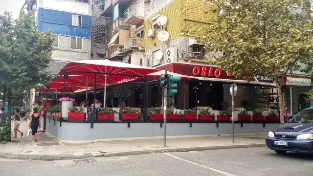 A street corner with red awnings and parasols, flower planters and apartment buildings behind