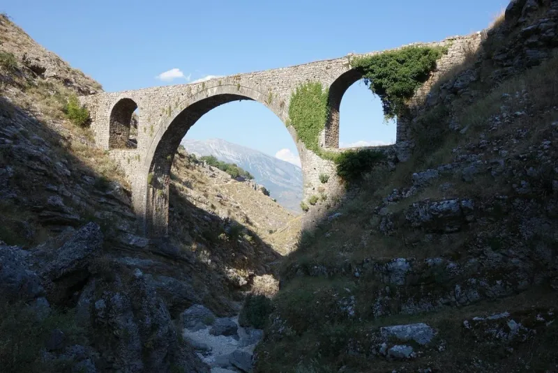 A stone arched bridge over a rocky gorge with a mountain peak framed inside the main arch