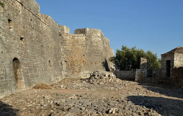 Massive stone fortress walls against a blue sky, with a courtyard strewn with fallen stones