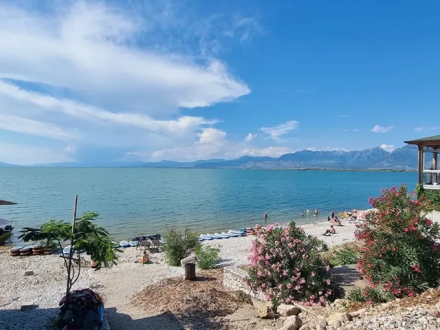 A wide lake with mountains on the horizon, kayaks on a pebble shore and flowering oleander shrubs