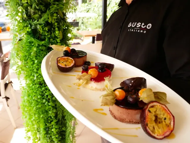 A waiter holding a long plate with mousse desserts, passion fruit and berries, beside a plant wall