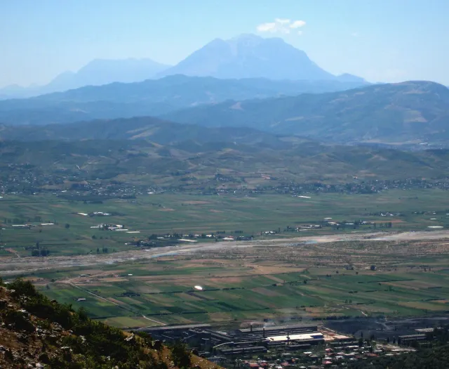 A wide valley with green fields and a settlement, and a mountain massif rising hazily behind