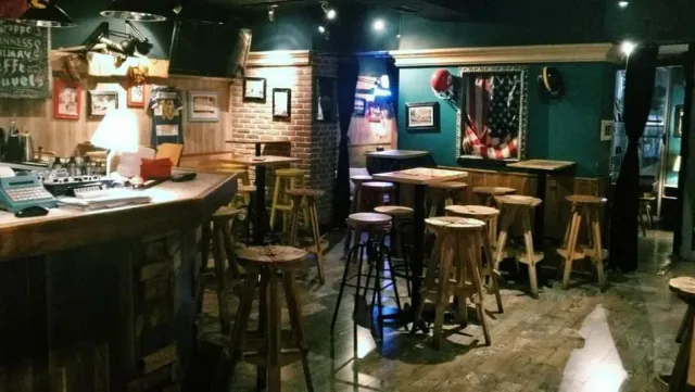A dim pub interior with a wooden bar, high stools, brick walls and framed pictures