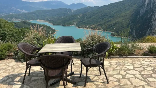 A table and wicker chairs on a stone terrace facing a turquoise lake among mountains