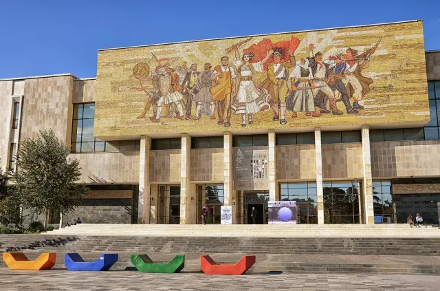 A huge mosaic of flag bearing figures above a modern museum facade on a paved plaza