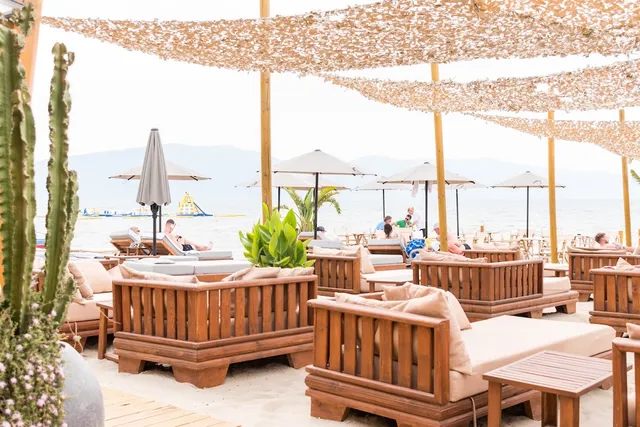 Wooden sofas with cushions on the sand under shade nets, and umbrellas facing the sea