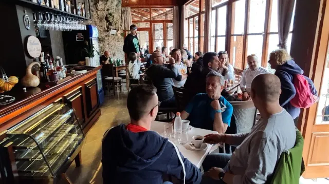 A hall crowded with diners around tables, a long bar counter and wooden windows in the background
