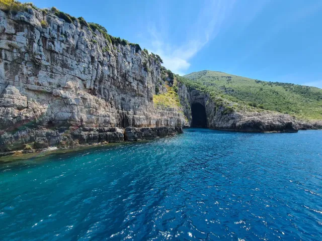 A high rock wall with a dark cave opening at the waterline and clear blue water