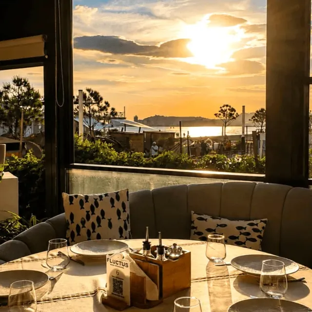 A set table by a large window with sunset over the sea, a sofa and cushions printed with fish