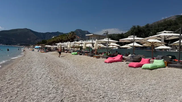 A white pebble beach with umbrellas and colorful bean bags, with green mountains in the background