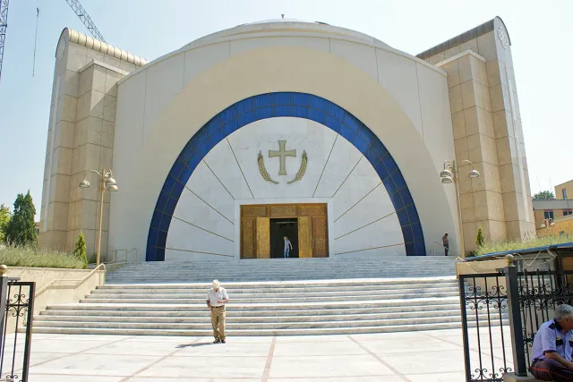 The white front of a modern cathedral with a blue arch and a gilded cross above broad steps