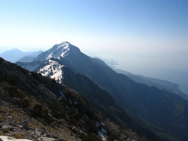 A snow capped summit above dark ridges and a bright strip of sea on the horizon