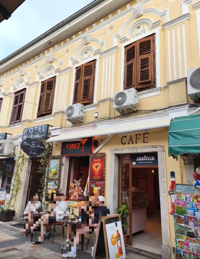A yellow building with wooden shutters and awnings, street tables and a postcard stand beside it