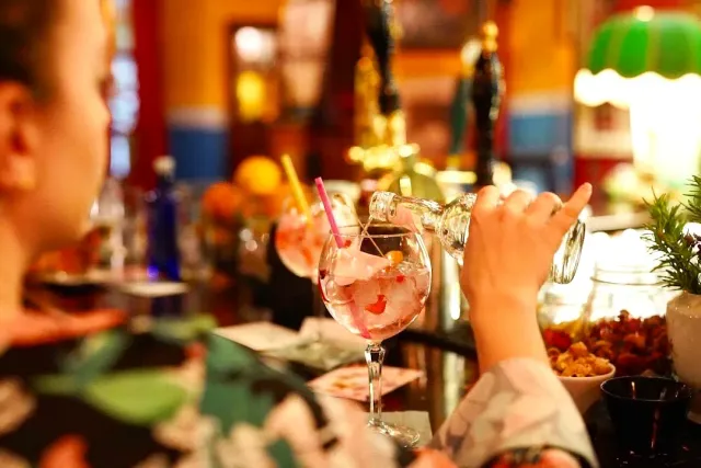 A hand pouring a drink into a cocktail glass with fruit on a bar counter lit in warm tones