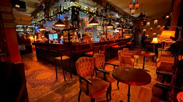 A long wooden bar loaded with bottles, upholstered armchairs and round tables in warm light