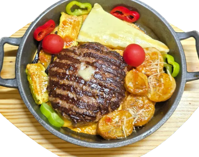 A grilled meat patty in a cast iron pan with potatoes, melted cheese and vegetables