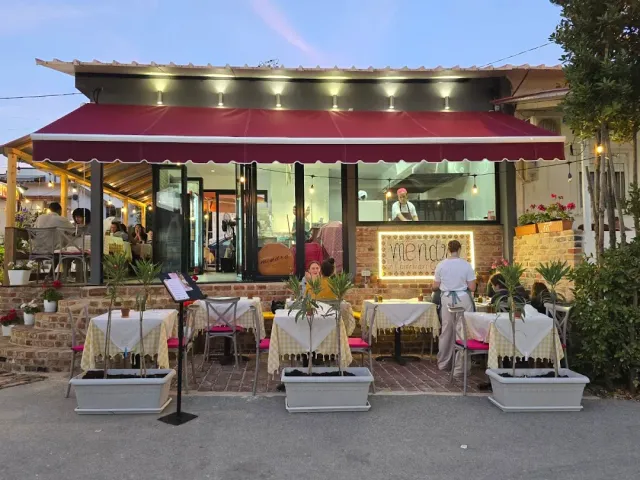 A restaurant front at dusk with a red awning, tables on the pavement and planters