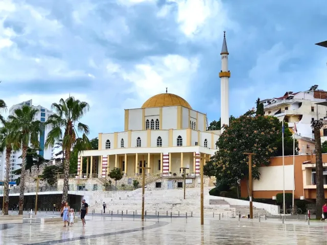 A white mosque with a golden dome and a tall minaret, palms and a wet square in front