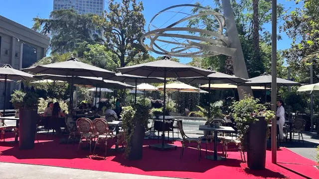 Tables under parasols on a red carpet in a city garden, with a ring-shaped metal sculpture above