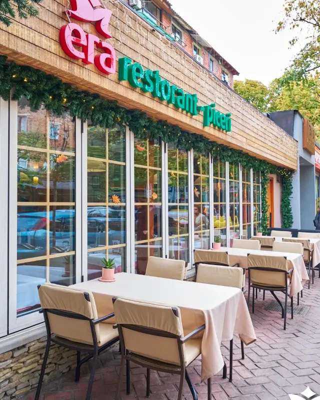 Outdoor tables with beige cloths facing a glass front trimmed with a green garland