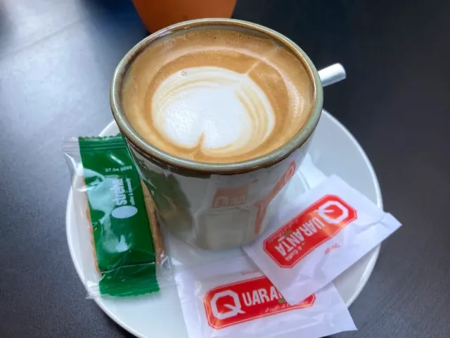 A cappuccino cup with foam art on a white saucer beside sugar packets and a biscuit