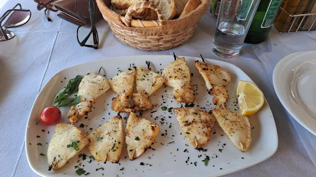 Grilled squid skewers with parsley and lemon on a white platter, with a bread basket in the background