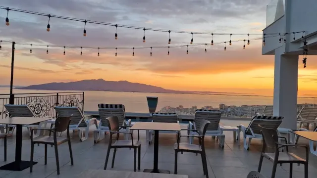 A terrace with a string of bulbs and plastic chairs, facing the sea and a mountain at orange sunset