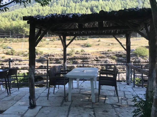 A wooden pergola with a white table and chairs on a stone terrace facing wooded hills