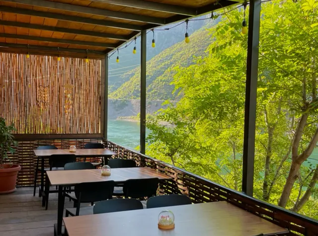 A covered terrace with dark tables, hanging bulbs and a view of a river and a green slope