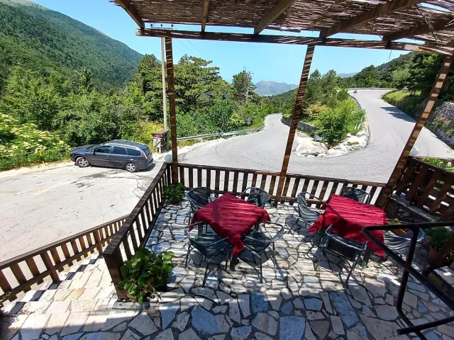 A stone terrace with red cloths under a wooden pergola, and a road winding among mountains