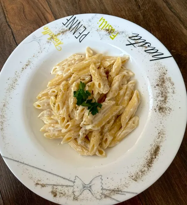 A plate of penne pasta in cream sauce with a parsley leaf in the centre on a wooden table