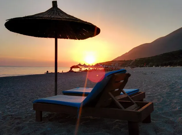 A straw parasol and a blue sun lounger on the beach facing a sunset and mountains