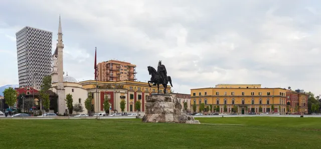 A bronze equestrian statue on a stone plinth at the centre of a square, facing yellow government buildings and a minaret