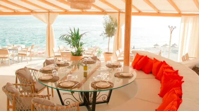 A glass table set under a pergola, a curved white bench with orange cushions and a sea view