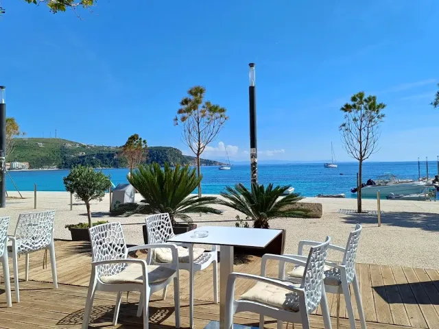 A white table and chairs on a wooden deck facing a blue bay, palms and boats