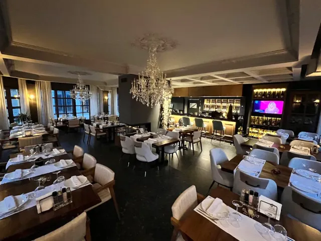 An elongated hall with crystal chandeliers, tables in white cloths and a lit wine bar behind